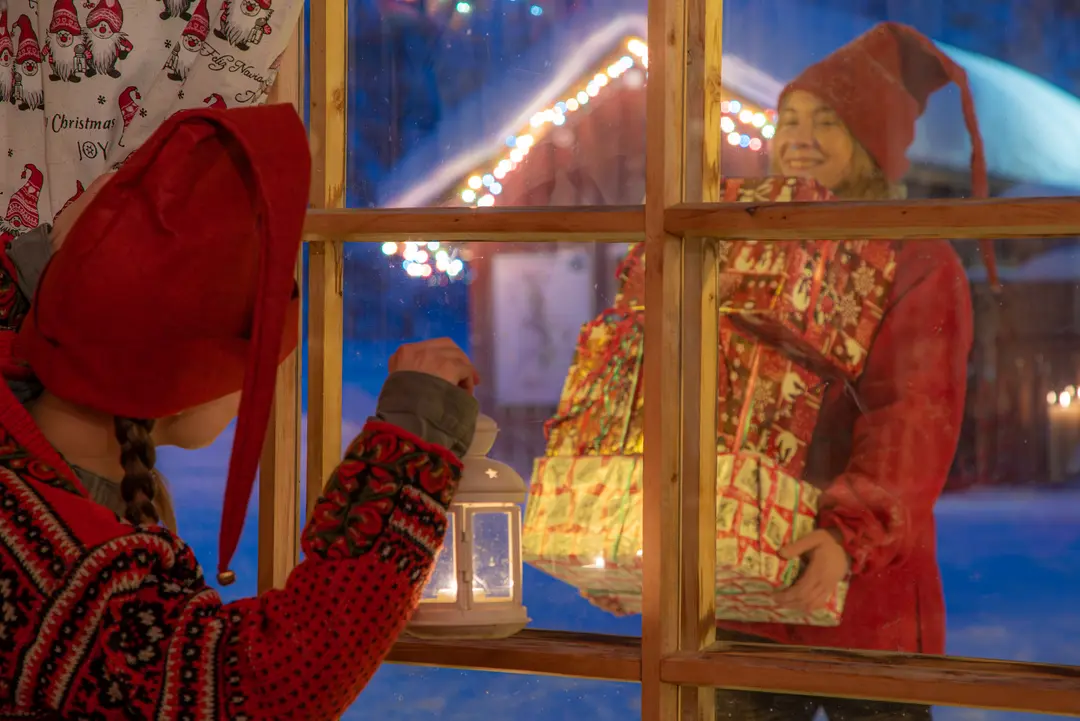 Child dressed as an elf peering through a frosty cabin window at another elf with Christmas presents
