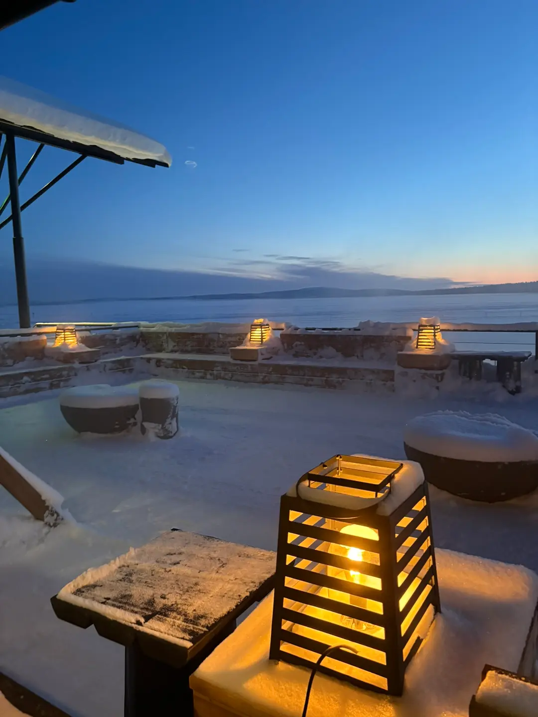 Snow-covered terrace with glowing lanterns overlooking a frozen lake at twilight