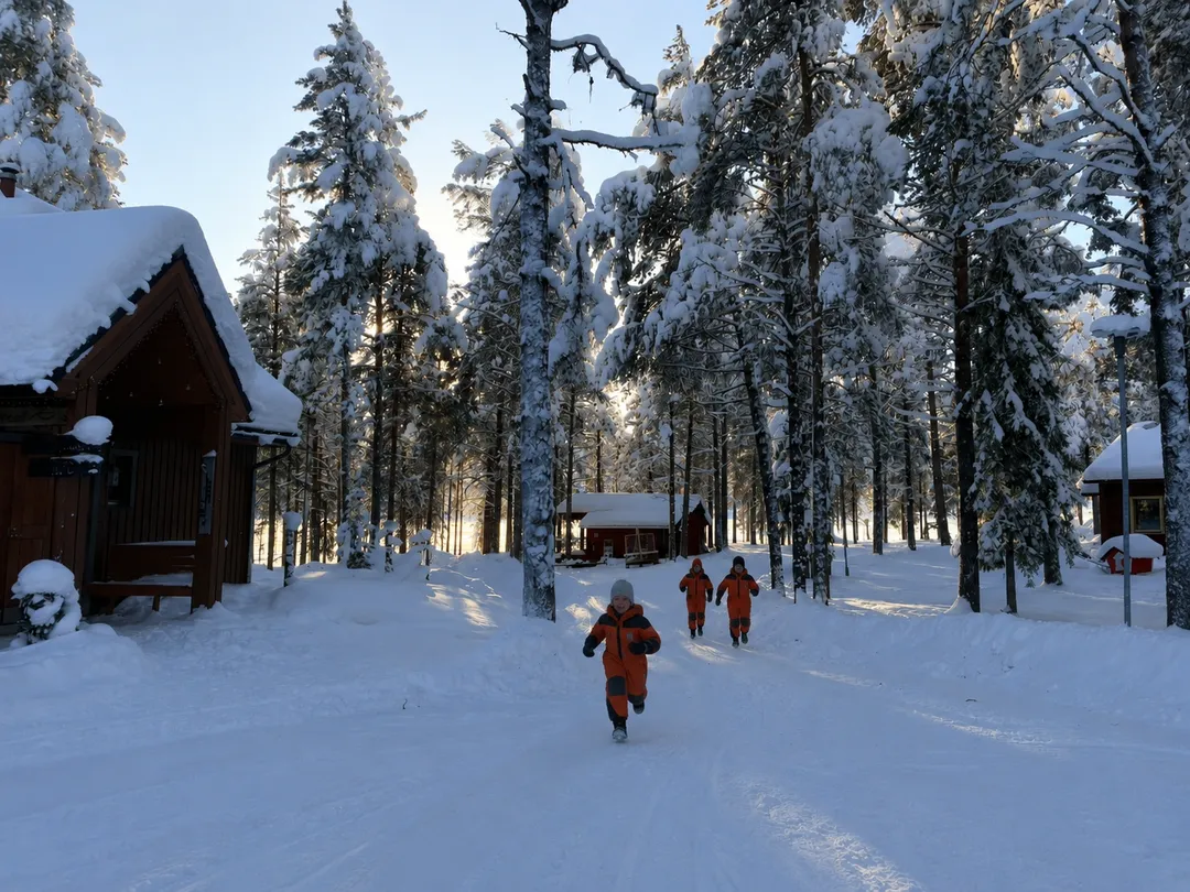 Family walking between snow-covered cabins and pine trees at Torassieppi